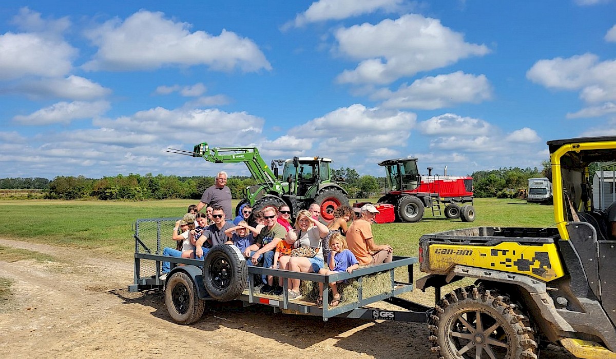 farm tour guests sitting on hay on a trailer being pulled by a side by side through farm fields